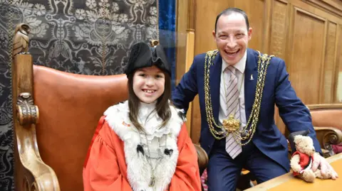 Leeds City Council A 10 year old girl with brown hair wears a red cape and a frilly white collar. A fur lines her cape and she wears a black ceremonial hat. She sits on a large brown leather chair with owls carved into the frame. Next to her a man in a blue suit smiles. He has dark hair and wears a large gold chain around his shoulders. In his hand is a teddy bear.