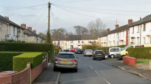 Google Google street view showing gardens and white houses lining the road with cars parked on both sides.