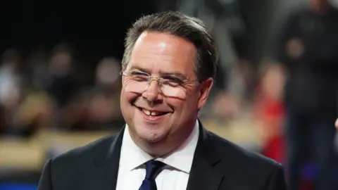PA Media Craig Hoy - a man with glasses, short dark hair and a suit - smiles while standing at an election count