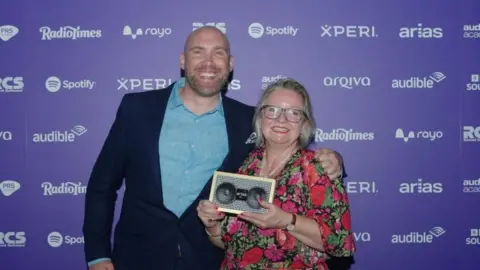Joe Sims, a bald man wearing a blue shirt and dark jacket, stands smiling next to Nicki Ledgard, a woman with blonde hair and glasses, who is holding a trophy that looks like an audio speaker.