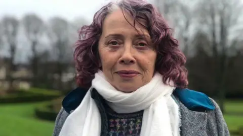 Margaret Martin is an older woman with curled, purple hair. She is looking at the camera with a neutral expression on her face. She is wearing a white scarf, a grey coat and a colourful jumper underneath. She is standing in the middle of a park, but the background is blurred.