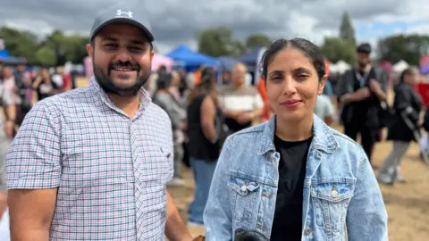 a man wearing a checked shirt and a baseball cap stands next to a woman with dark hair and a light blue denim jacket