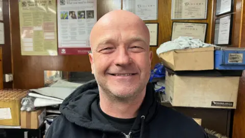 John Devine/BBC Martyn Hopkins smiling at the camera in his office. He has a bald head and stubble on his face. He is wearing a black hoodie. Behind him are various boxes and certificates.