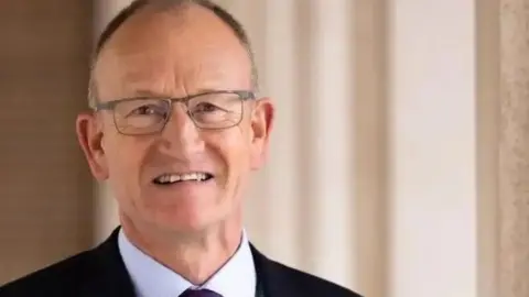 Hertfordshire County Council Richard Roberts with short dark hair and silver-framed glasses wearing a black jacket, purple shirt and dark purple tie. He is standing in front of brown curtains.