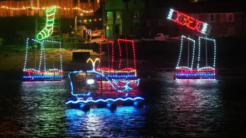 A picture of the lights in Mousehole. There is three boats with lights on pictured. One is displaying a stocking, one has a whale on it and one has a cracker. They are all lit up in different colours.