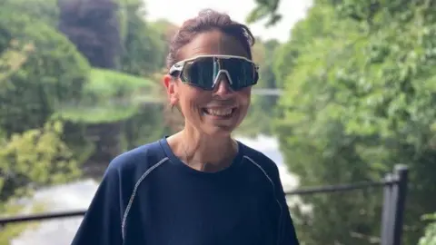 Sally Hurst/BBC Hannah Corne, smiling and wearing dark sunglasses and a navy T-shirt. In the background are railings with a river running behind. The area is surrounded by greenery.
