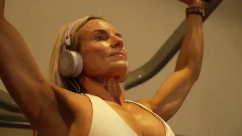 Jane Woodhead has blonde hair and is wearing a white sports top and headphones while using a weight machine in a gym.