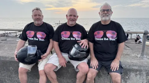 Zoie O'Brien/BBC A group of men sitting in a line on a concrete sea defence wall, the grey sea and horizon behind them. They are all wearing shorts black tshirts with chase the sun written in white across the chest and an image of a pair of sunglasses with palm trees reflected in them. They all have grey hair and beards. The two men to the left have black motorcycle helmets held down on their laps