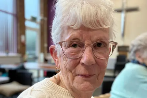 A woman with white hair, who is wearing glasses and a cream jumper, smiles as she looks into the camera while sitting in a community hall with large windows.