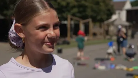 A girl in a pink T-shirt is seen from her shoulders up smiling.