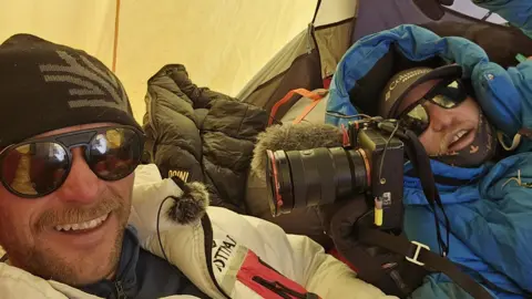 Tim Howell Two men in jackets and sunglasses are lying down in a tent. One of the men is holding a camera.