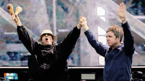 Big Brother Recordings Liam and Noel Gallagher with their hands in the air as they walk on stage. Liam is wearing a beige bucket hat and a brown jacket and holding a pair of wooden maracas in the right hand. Noel is wearing a blue blazer-style jacket.