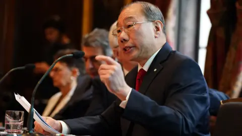 PA Media Dr Shoo Lee, who has thinning black hair and wears rectangular-framed spectacles and a navy-blue suit over a white shirt, speaks into a microphone while raising his left hand. 