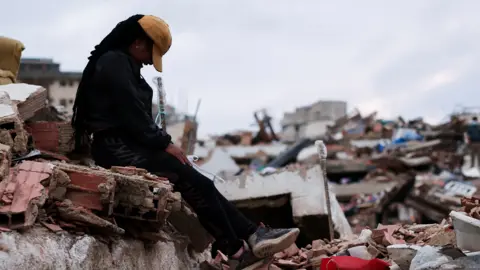 Reuters Hilyerin Subero, 28, reacts after finding her husband Jeison Nieto’s keys, in the aftermath of the deadly June 24 earthquakes, in La Guaira, Venezuela, July 6, 2026. She is sitting on a mound of rubble.