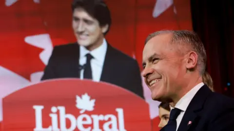 Mark Carney smiles in the foreground while Justin Trudeau's speech is beamed onto a screen in the background