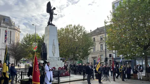 An Armistice Day service is held at the War Memorial in Leeds. Military flags are held aloft by service personnel as a vicar stands at the front of the monument. Crowds line the side.