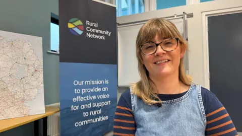 Emma McAleer tiene el pelo rubio hasta los hombros y lleva gafas. Lleva un vestido vaquero azul sobre una blusa azul oscuro con finas rayas naranjas en las mangas. Detrás de ella hay una pancarta desplegable con fondo azul y letras blancas que dice: Red Comunitaria Rural. Nuestra misión es dar voz y apoyo efectivos a las comunidades rurales.