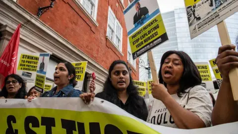 Getty Images A row of striking nurses stand in their picket line at Guy's & St Thomas' Hospital, in London. Dressed in off-duty clothing, four women can be seen behind a yellow and white banner which spells out part of the hospital's name. One of the women is holding a yellow and black placard with the slogan "stop exploiting your nurses".