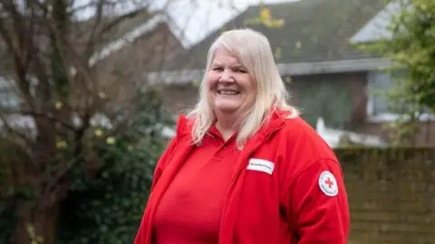 British Red Cross A woman with white-blonde hair standing outside wearing a red polo shirt and a red fleece with the British Red Cross logo on the arm.