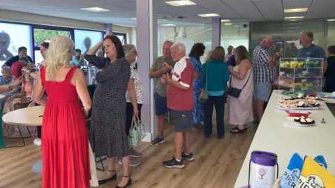 Visitors mingle in a brightly lit community centre with purple pillars and wooden flooring. Tables and chairs are arranged throughout the space, with a refreshments table on the right offering cakes, snacks, and a donation tin. The setting is informal and sociable
