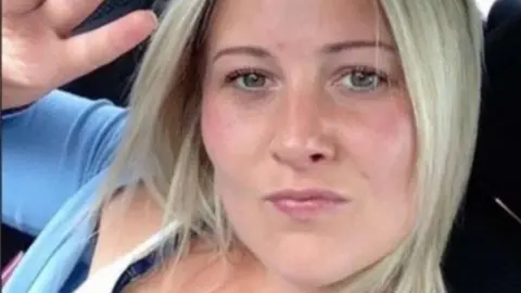 Joanne Penney seen in selfie taken sitting in a car. She is making the peace sign hand gesture and has shoulder-length straight blonde hair and wears a light blue cardigan.