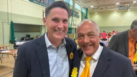 Charlie Maynard and Andy Graham - both wearing suits. Andy Graham has a yellow tie and a yellow rosette. Behind them is the election count underway.