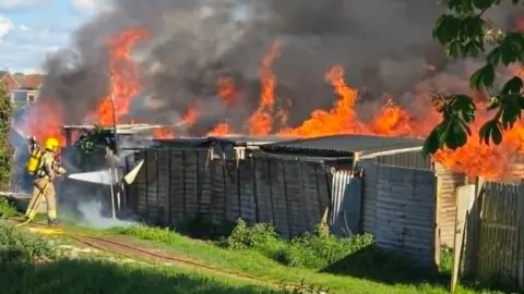 A number of sheds at North Lynn allotment in King's Lynn. There is a firefighter with a house at the back of the land. There are large orange flames and smoke trailing into the sky from the blaze.