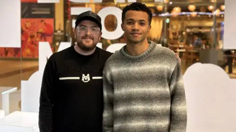 Two men stand side by side smiling at the camera, with one wearing a black Moncler sweatshirt and cap and the other in a grey gradient knitted jumper. They are standing in front of a glass window with large white letters partially visible behind them. A restaurant with warm lighting can be seen through the glass in the background.