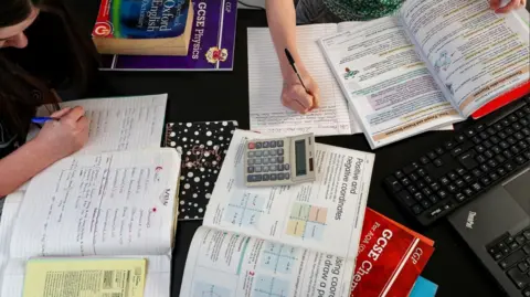 PA Media Looking down at a number of open text books, note books, a keyboard and a calculator on a table with two people's hands writing in books.