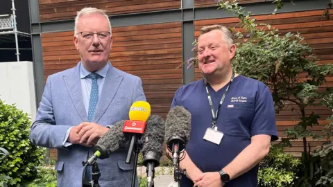 The health minister and Mr Taylor giving a press conference. Nesbitt, left, is wearing a light grey suit and blue tie. He has glasses and short grey hair. Mr Taylor, right, is wearing navy scrubs and has a lanyard around his neck. He has short greyish hair.