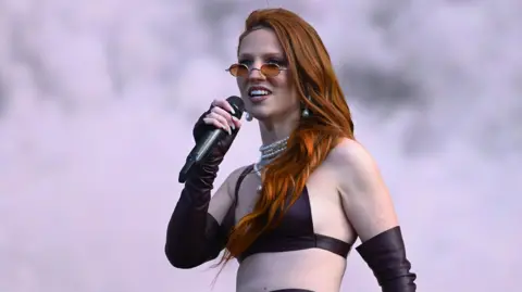 Jess Glynne, wearing sunglasses and a bikini-style top, singing into a microphone while on stage