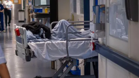 A hospital bed halfway out of a ward doorway. Someone is lying in the bed covered by blue and black blankets. A corridor is on the left with doctors and staff walking down it. 