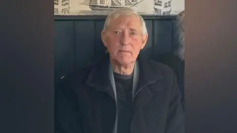 An older man wearing dark coat. He appears to be sitting on a wooden-backed bench, possibly in a pub or restaurant.