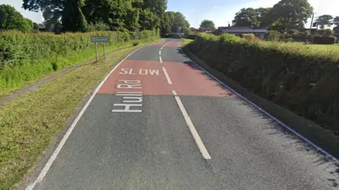 A street view of the B1238. There is a sign up ahead on the left that says Albrough, please drive carefully and the road is lined on either side by grass verges and shrubs.