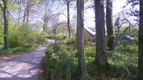 Google Google Street View of a winding narrow lane through a wooded area. Through the trees is a barn and a blue van.