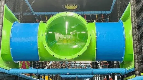 The image shows a section of a children's indoor playground. It shows a large cylindrical tunnel made of blue and green plastic, with a transparent green middle section. The tunnel is supported by blue padded bars and enclosed with safety netting. 