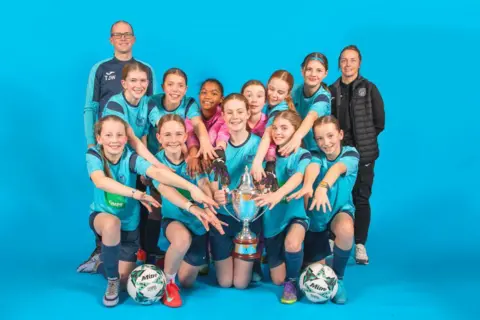 Stephen Tierney All the girls are smiling and reaching out to touch the cup. There are two footballs in the foreground.