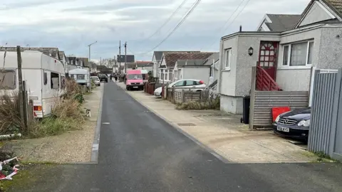 RICHARD KNIGHTS/BBC Hillman Avenue in Jaywick in 2024. The road shows prefabricated buildings of non standard construction both sides. Small caravans and vans are parked outside some of the houses, you can also see overgrown grass to the front of one of the properties