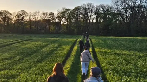 Lauren Murphy A group of walkers crossing a ploughed grass field with their backs to the camera at sunset