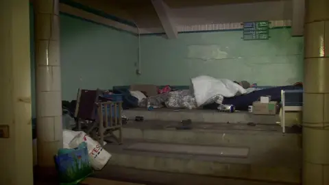 A person sleeping rough. There are blankets and plastic bags everywhere.