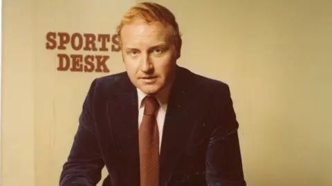 John Bennett pictured in a suit, sat at a brown desk with Sports Desk sign on wall behind him