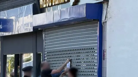 Image shows enforcement officers putting a closure order notice on the metal grilles on the outside of the West Kirby Vape Shop