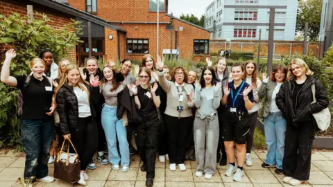 Construction Anglia The seventeen female students from West Suffolk College are pictured. They are in a close group facing the camera. They all have their hands up celebrating with smiles on their face. Buildings from the college can be seen behind them.