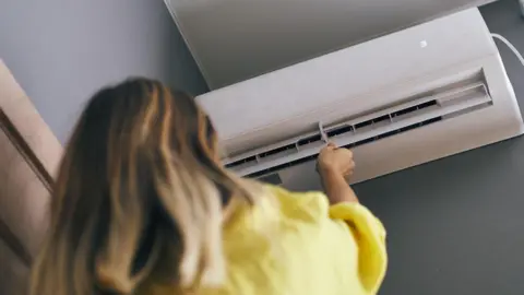 A woman with brown hair is wearing a yellow jumper and is pointing a control at a white air conditioned unit on a wall.