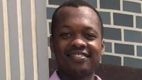 Patrice Che - a man with short black hair wearing a pink shirt and smiling. He is standing in front of a grey brick wall.
