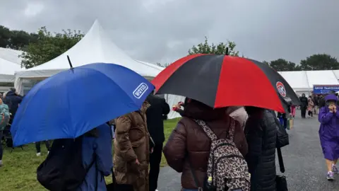 A group of people stand and walk along a pathway holding open umbrellas on a grey, rainy day, with large white event tents set up in the background. The scene looks like an outdoor show or fair, with crowds moving between stalls while sheltering from the rain.