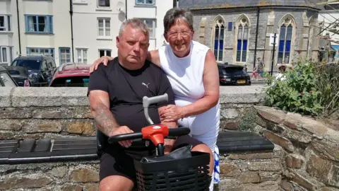 Handout Wendy and Paul posing in front of a stone wall that has a road with cars, some houses and a church-style building behind them. Paul is wearing a black T-shirt and is sat on a black mobility scooter and holding his walking stick. Wendy is wearing a white T-shirt and trousers and is stood next to to him with her left arm on his shoulder.