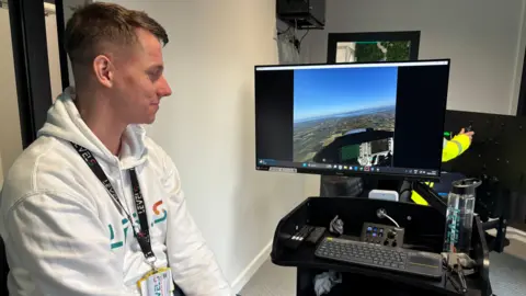 A blond-haired man in a white hoodie is looking at a computer screen. 