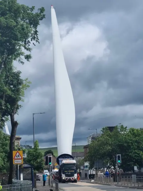 CWP Energy A large turbine blade on the back of a specialist lifter passing through a town. There are traffic lights and a sign for a school crossing patrol at the side of the road and a few people appear to have gathered to watch