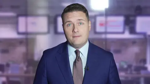 Wes Streeting, he has short dark hair, is clean shaven and is wearing a blue suit with white shirt and a metallic purple-grey colour tie